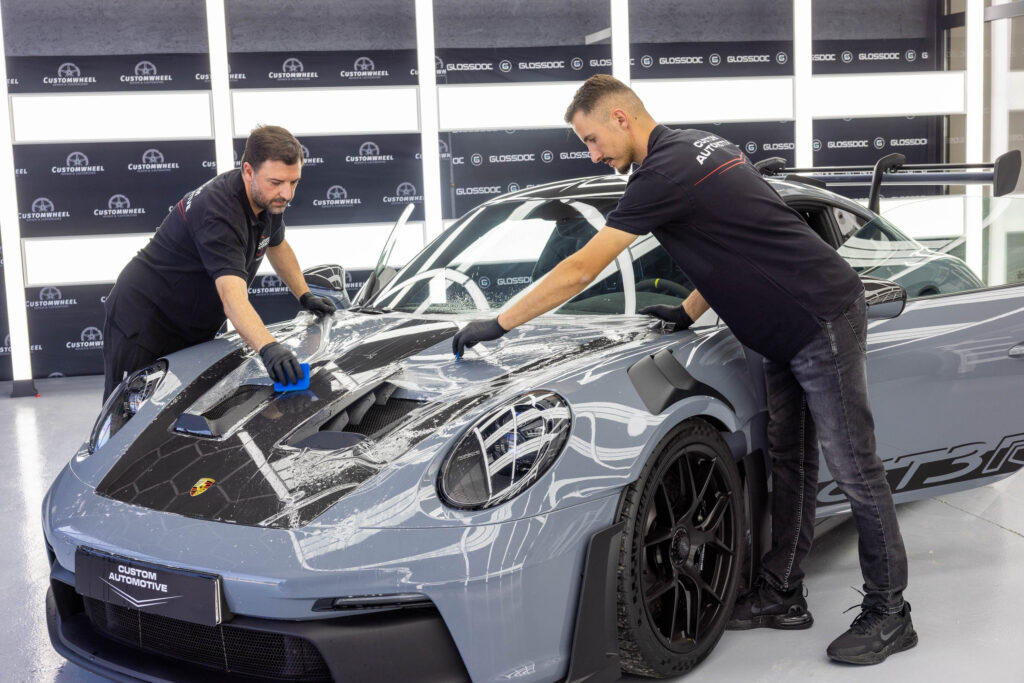 Aufziehen von PPF Schutzfolie auf Motorhaube von grauem Porsche GT3 RS durch zwei maennliche Facharbeiter